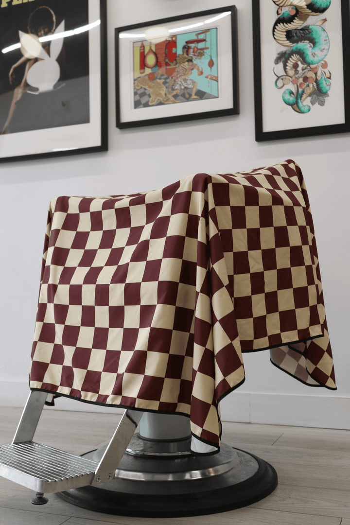 Low-angle view of Check Cape draped over barber chair. Showcasing checker pattern with burgundy and tan colours.