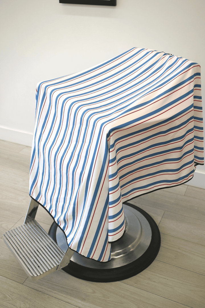 A traditional barber cape with white, blue and red stripes. Side view of Blood and Bandages Cape draped over barber chair.