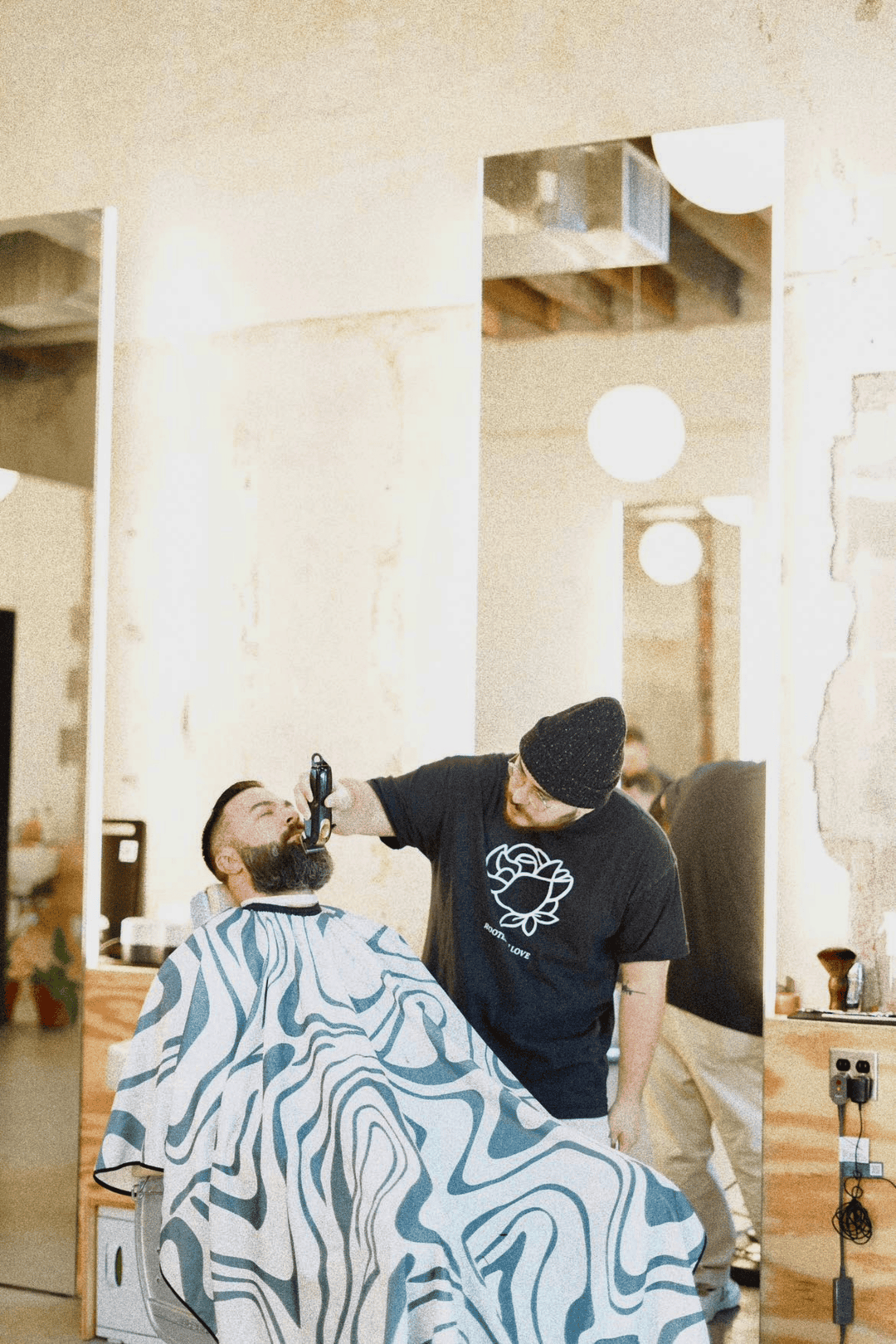 A barber trimming the beard of a customer wearing the Aurora Barber Cape by High Top Capes
